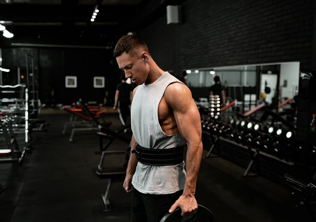 Homme musclé en pleine séance de musculation dans une salle de sport, portant une ceinture lombaire et tenant un disque de poids.