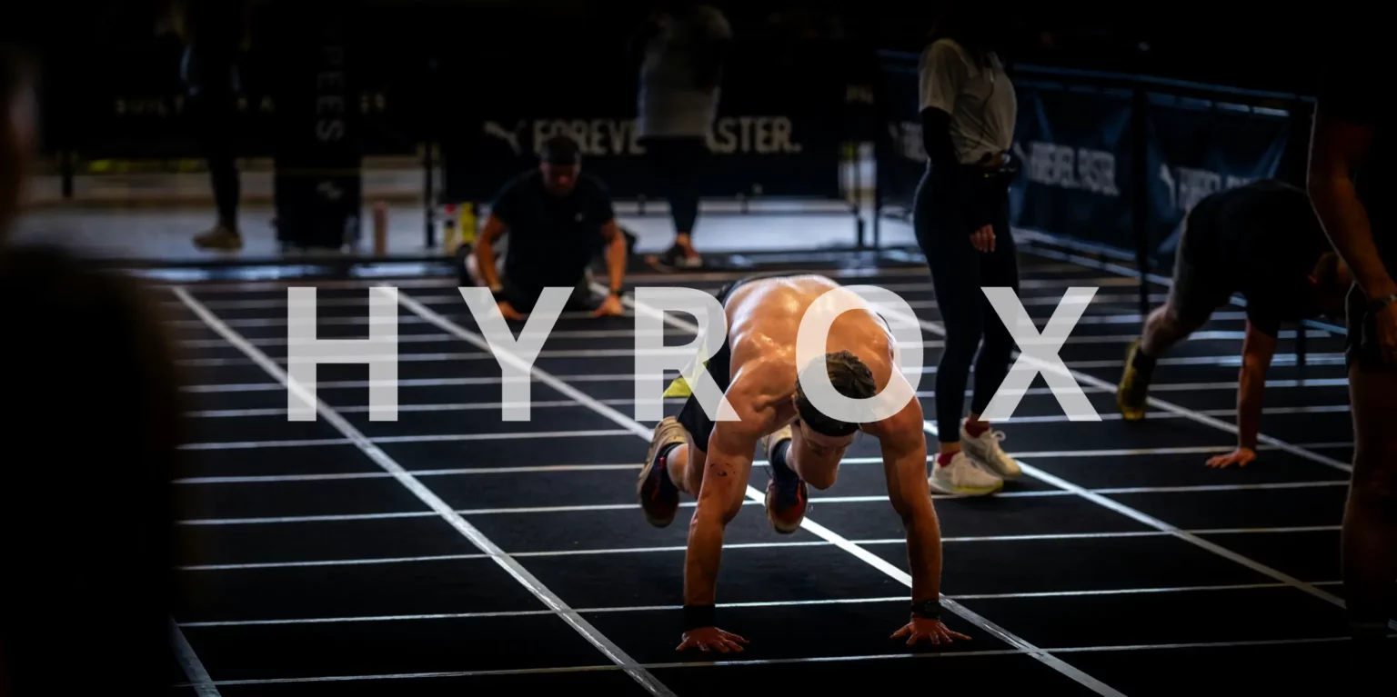 Athlète en pleine épreuve Hyrox réalisant un exercice fonctionnel sur tapis de compétition, mettant en avant l’endurance, la force et la préparation physique typiques de ce format d’entraînement hybride.