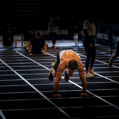Athlète réalisant des burpees dans une zone de compétition HYROX, torse nu et en pleine phase dynamique de l'exercice sur un sol quadrillé.
