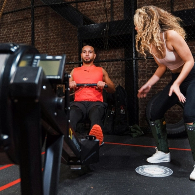 Homme utilisant un rameur en salle de sport, accompagné d’une coach, dans un espace d’entraînement fonctionnel aux murs en briques.