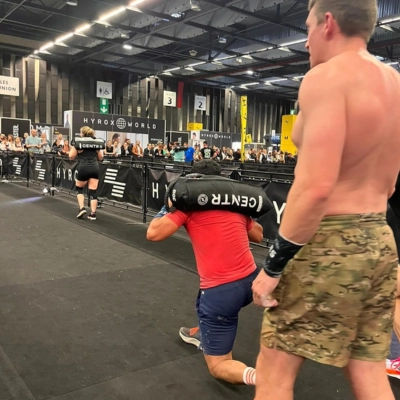 Athlète réalisant des sand bag lunges lors d'une compétition HYROX, sac lesté sur les épaules, devant un public rassemblé dans une salle dédiée a l'HYROX