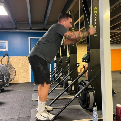 Athlète utilisant un SkiERG lors d'un entrainement HYROX, concentré sur le mouvement de tirage vertical, entouré d'autres compétiteurs.