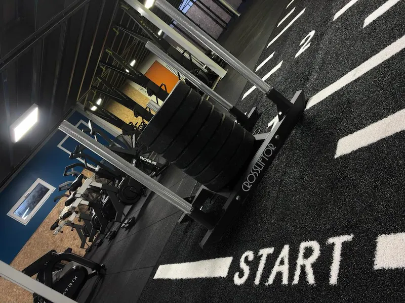 Tapis HYROX de sprint en salle d'entraînement, avec une luge de poussée CROSSLIFTOR chargée de disques, positionnée sur la ligne de départ.
