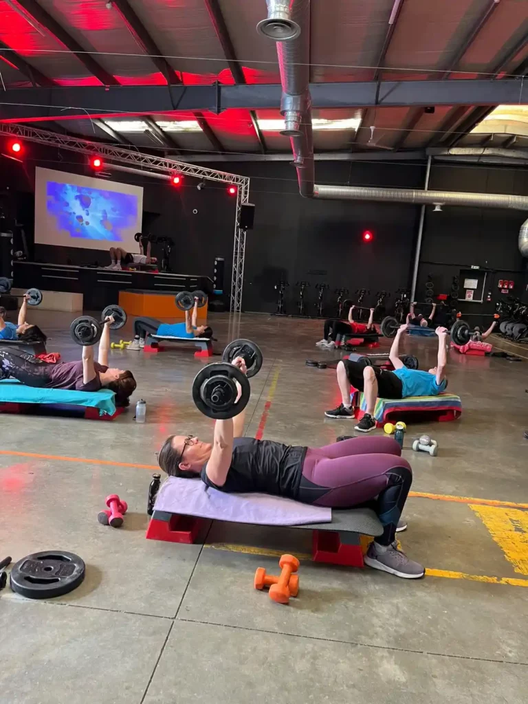 Cours de bodypump à Beynost avec participants en pleine séance de musculation sur step, effectuant un développé couché avec barre dans une salle de fitness équipée.